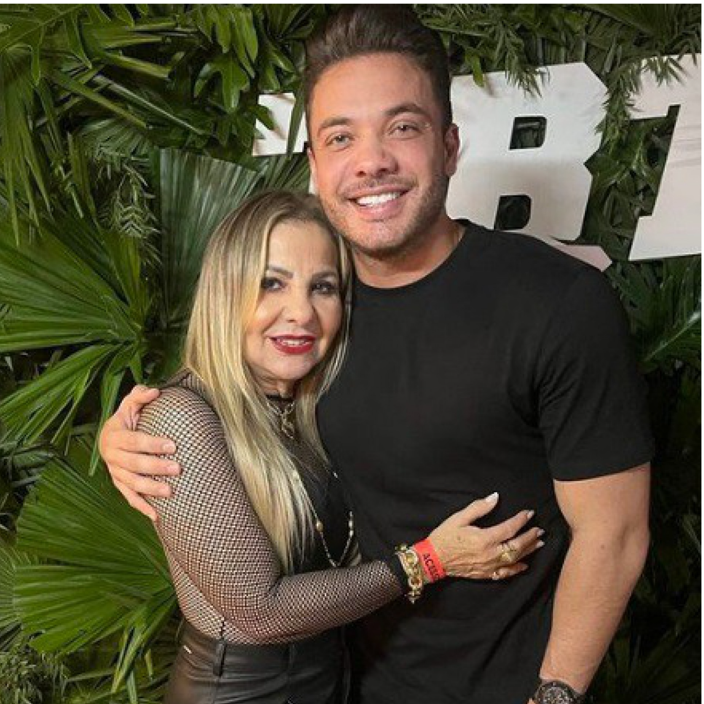 Dona Bill, mãe de Wesley, ao lado de Wesley. Estão abraçados. Ao fundo, algumas plantas, o chão é vermelho. Os dois estão sorrindo. Wesley usa uma camisa preta e uma calça preta, além de um relógio no pulso esquerdo. 