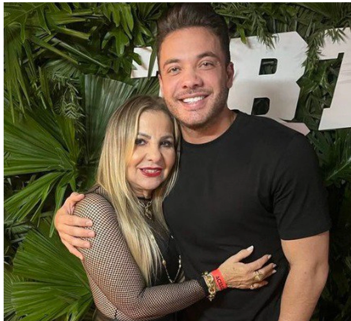 Dona Bill, mãe de Wesley, ao lado de Wesley. Estão abraçados. Ao fundo, algumas plantas, o chão é vermelho. Os dois estão sorrindo. Wesley usa uma camisa preta e uma calça preta, além de um relógio no pulso esquerdo. 