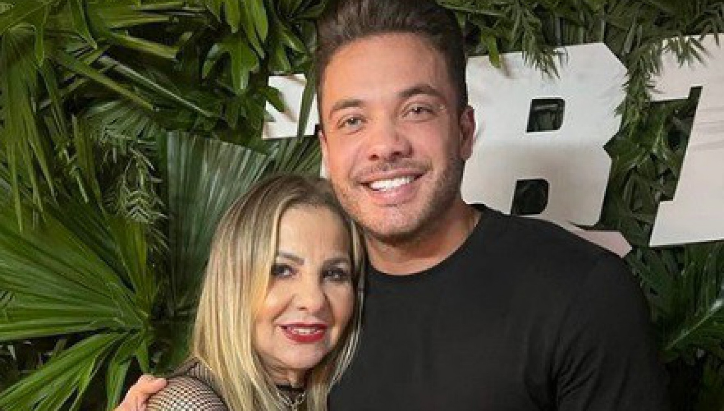 Dona Bill, mãe de Wesley, ao lado de Wesley. Estão abraçados. Ao fundo, algumas plantas, o chão é vermelho. Os dois estão sorrindo. Wesley usa uma camisa preta e uma calça preta, além de um relógio no pulso esquerdo. 